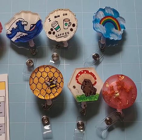 These handmade badge reels feature unique resin art pieces on retractable clips, including designs like a wave, coffee theme, rainbow, honeycomb with a bee, and a sloth. Each reel is detailed and colorful, adding a personalized touch to ID badges or keys.