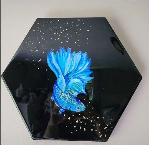 This handmade resin and acrylic decoration features a vibrant blue betta fish on a glossy black hexagonal background. Glittering accents and scattered specks enhance the design, creating a dynamic and eye-catching piece.